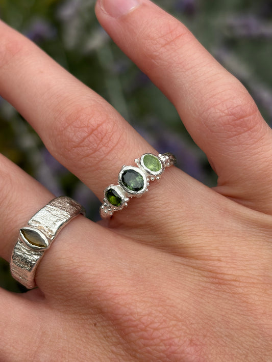 A close-up of a hand wearing two silver rings. The primary product shown is a delicate silver band ring with three oval green gemstones