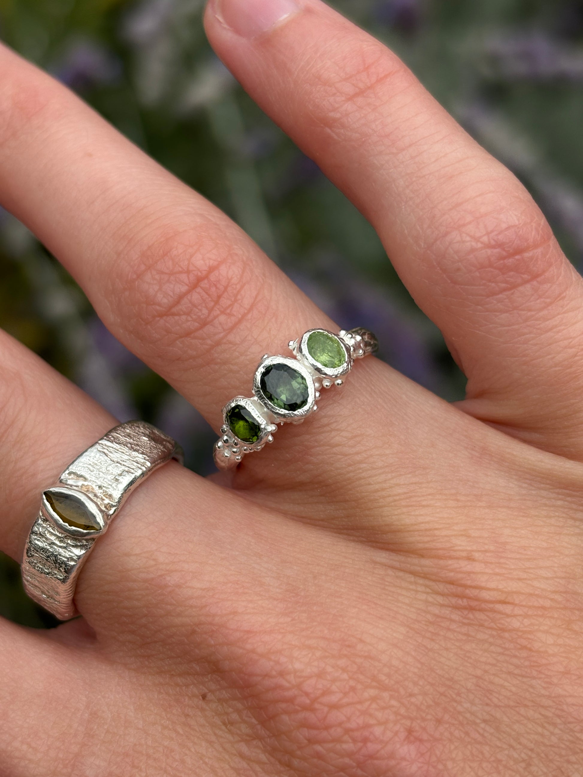 A close-up of a hand wearing two silver rings. The primary product shown is a delicate silver band ring with three oval green gemstones
